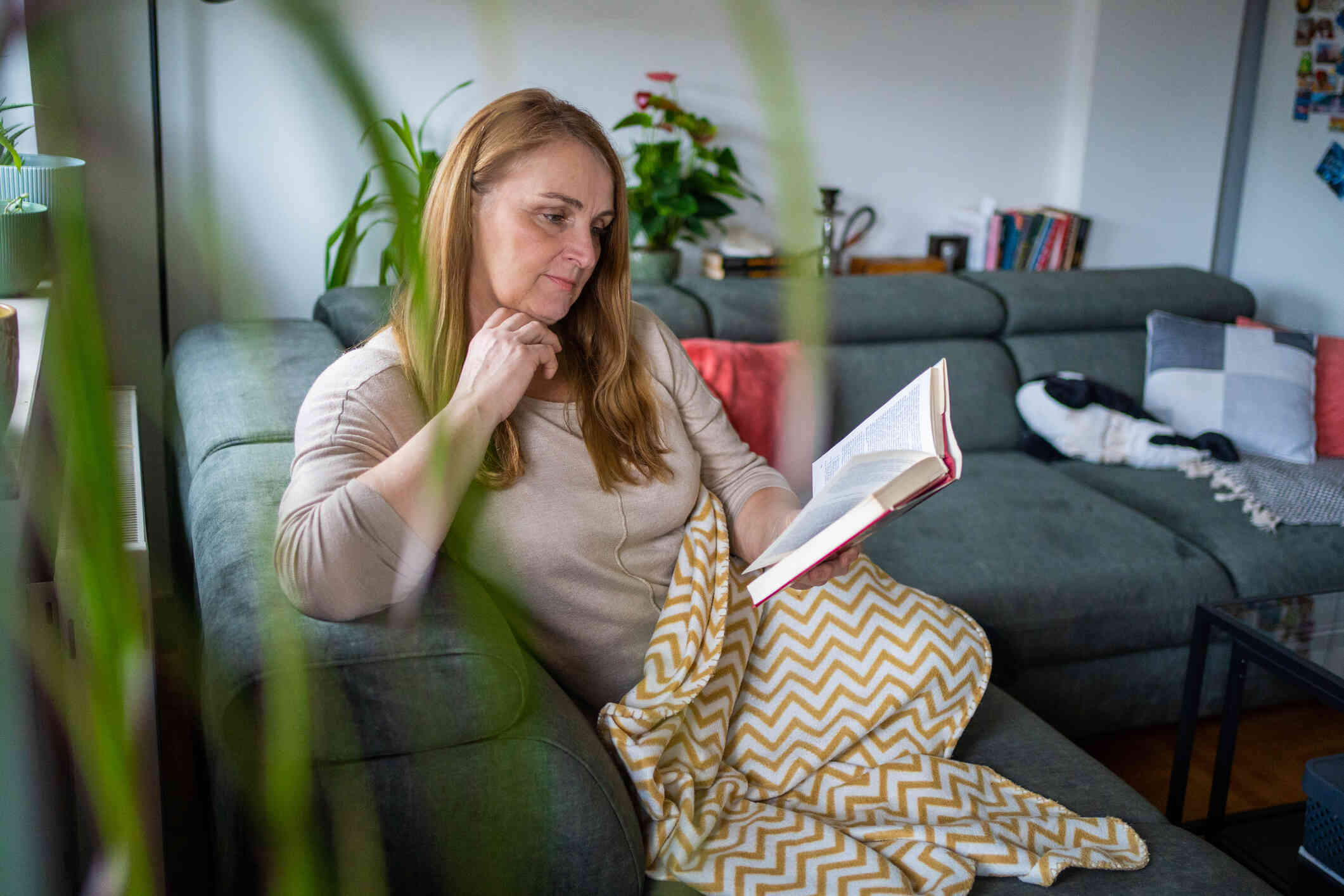 A middle aged woman sits under a blanket on the couch while reading a book.